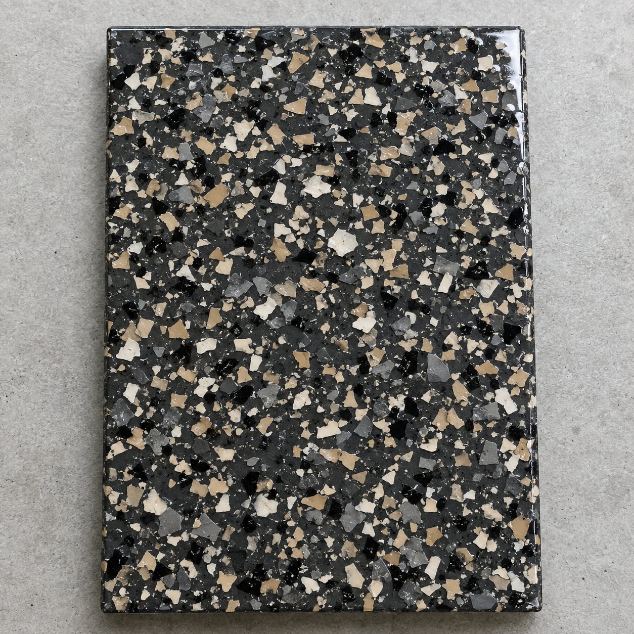 A broadcast vinyl chip flake concrete floor coating sample in charcoal, tan, cream, and black.